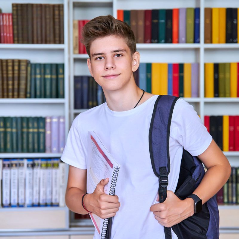 Portrait of male teenage student looking into the camera in library Portrait of male teenage student looking into the camera in library. Smiling positive guy 16, 17 years old with backpack, notebooks. Education, high school, college, adolescence concept